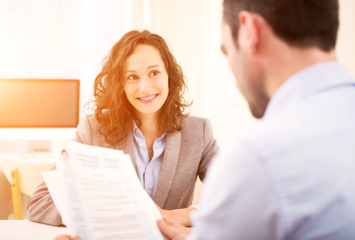 Frau mit lockigen Haaren und beigem Blazer sitzt einem Mann im hellblauen Hemd gegenüber, der sich ihren Lebenslauf ansieht